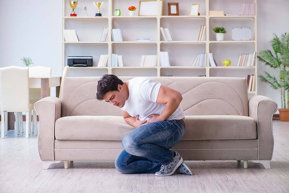 Young man kneeling by couch clutching stomach in pain, in living room with bookshelves|Man passed out on table with bottle of alcohol nearby, depicting the harmful effects of alcohol addiction and the need for intervention|Person collapsed on floor from suspected alcohol poisoning, with empty bottles nearby and someone checking on them