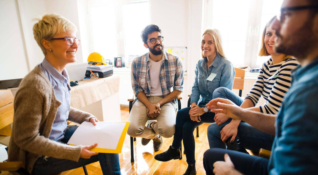 Diverse group of people sitting in a circle during a therapy session at an addiction treatment center, led by a female therapist|Female therapist in white top sitting in a green armchair, taking notes while listening to a patient in a yellow top, in a cozy office setting with bookshelves and plants|Bright and inviting rehab facility bedroom with wooden bed, green walls, skylight, floral pillows, and modern amenities including TV and bedside lamps