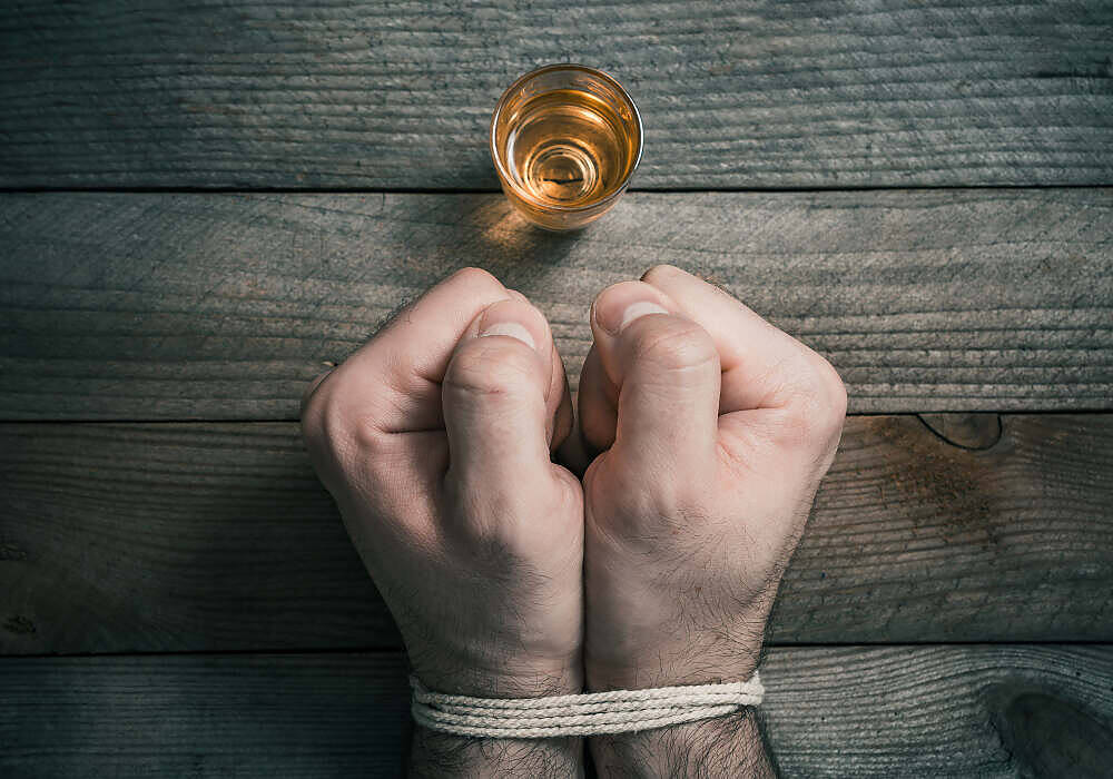 Clenched fists tied with rope next to a glass of alcohol on a wooden surface, symbolizing addiction struggles|Man sitting on a couch, holding a beer bottle, looking distressed, symbolizing alcohol addiction and its emotional toll.|Black and white image of a bearded man hunched over a bar counter, holding an empty beer glass, depicting the loneliness and despair of alcohol addiction