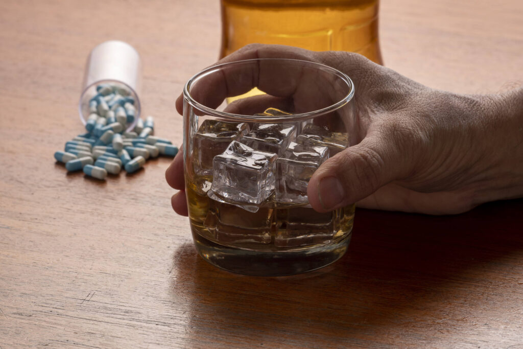 Glass of alcohol with ice cubes next to spilled antibiotic pills, illustrating the risks of combining alcohol and antibiotics|Hand holding glass of whiskey with ice cubes next to spilled blue antibiotic pills, illustrating the dangerous combination of alcohol and medication|Close-up of blue and white pharmaceutical capsules on a blister pack|Close-up of yellow prescription medication bottle with warning label about alcohol consumption, surrounded by other medication containers|Person in orange sweater pouring liquid medication from a bottle into a small measuring cup, demonstrating proper dosage measurement|Woman in grey sweater pouring liquid medication from a brown bottle into a measuring cup, highlighting the importance of proper dosing when taking antibiotics|Close-up of a prescription bottle label warning against mixing alcohol with medication, highlighting drowsiness and safety risks