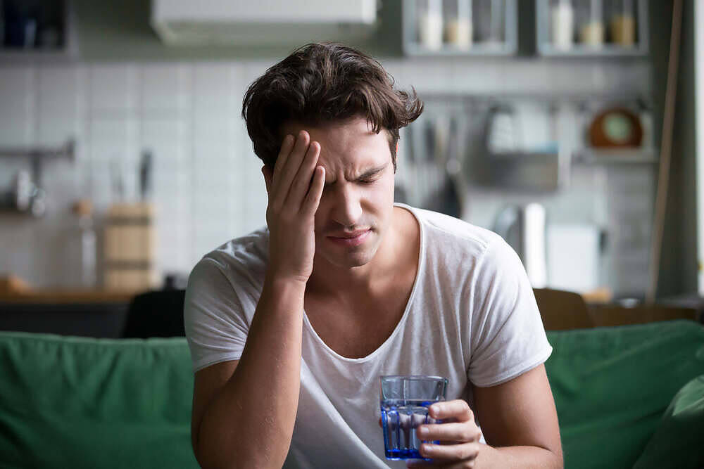 A man holding his head in pain while sitting on a couch, experiencing an alcohol-induced headache, and holding a glass of water|Woman lying in bed with eyes closed, touching her forehead, experiencing discomfort likely from an alcohol-related headache|Woman in grey suit holding her side with a glowing illustration of a liver, depicting alcohol-related liver pain and damage|Young man in white shirt and jeans sitting outdoors with head in hand, surrounded by bokeh light from trees, depicting struggle and contemplation in addiction recovery||