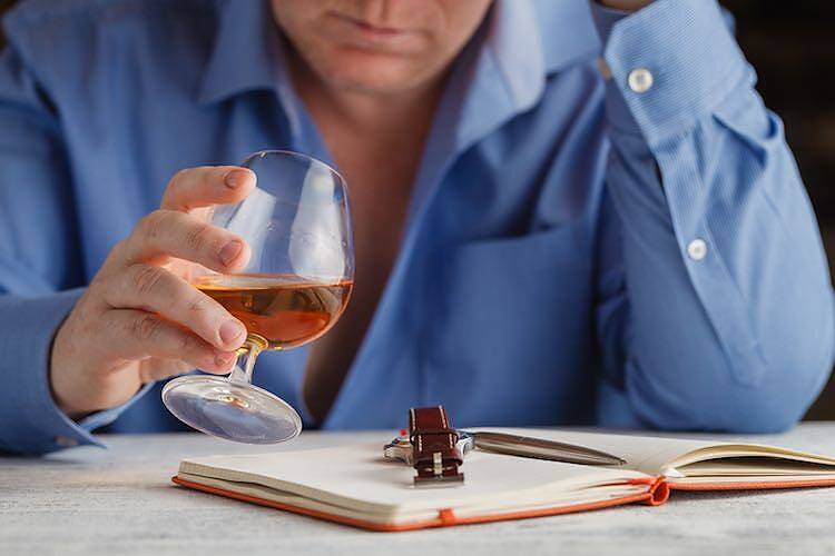 Person in blue shirt holding a glass of alcohol next to an open notebook, depicting work-related stress and alcohol use|Infographic showing government alcohol guidelines: 14 units per week limit, equivalent to 6 glasses of wine, 6 pints of beer or cider, or 14 shots of spirits