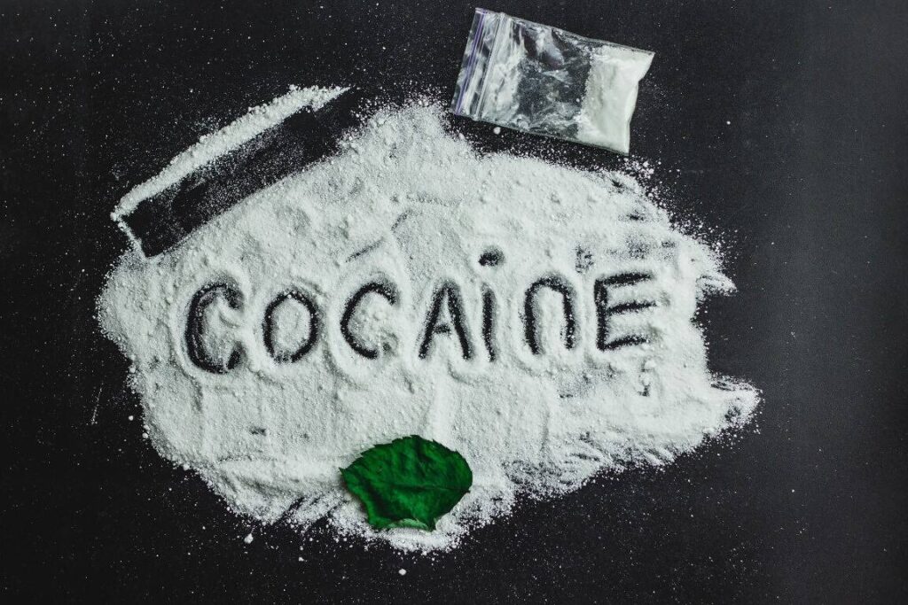 White powder spelling out 'COCAINE' on a black surface, with a small plastic bag and a green leaf, illustrating the illicit nature and health risks of cocaine use|Man covering face with hand as his body appears to disintegrate, illustrating the devastating physical and psychological effects of cocaine addiction