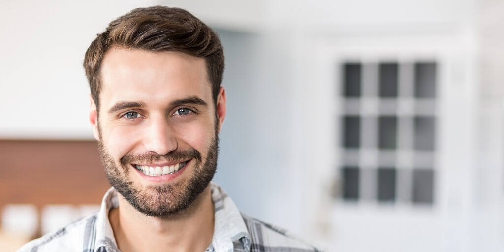 Headshot of a smiling bearded man with brown hair in a casual shirt, standing in a bright office space|Breathtaking East Midlands landscape with a winding river, lush forests, and wildflower meadows under a dramatic sky, symbolizing hope and renewal in addiction recovery|Portrait of a smiling young man with beard wearing a plaid shirt, representing positivity and success in addiction recovery|Collage showing a welcoming rehab facility exterior, cozy bedroom, and fitness area with people exercising, representing the comprehensive treatment approach in East Midlands rehab centers