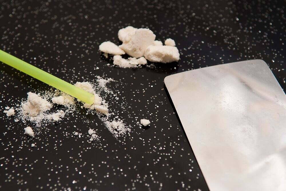 Close-up of crack cocaine rocks, powdered residue, green straw, and metal card on black background|A person in a grey hoodie and jeans sitting on the ground against a rusty wall, preparing to use drugs, illustrating the isolating and destructive nature of crack cocaine addiction