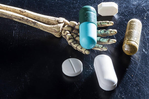 Skeletal hand holding colorful pills and capsules, illustrating the dangers of drug abuse and addiction|Close-up of white capsule pills with skull and crossbones symbol printed on them, symbolizing dangerous or addictive prescription medications on blue background|Stark image of a skeletal hand next to various prescription pills and capsules, symbolizing the lethal dangers of drug abuse and addiction|Two plastic bags containing dangerous drugs - one filled with blue pills, likely ecstasy or fentanyl, and another with white powder, possibly cocaine or methamphetamine|Prescription bottles of Oxycodone with white pills spilled out, illustrating the potential for abuse of dangerous prescription drugs|Hands rolling a marijuana joint with dried cannabis in a plastic bag, illustrating potential dangers of drug use