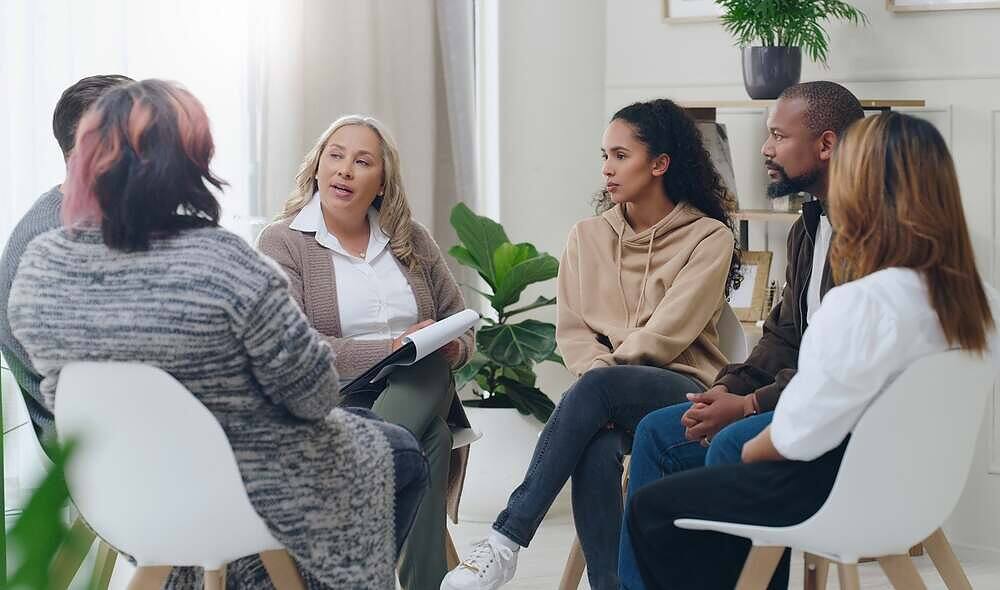 Therapist leading a diverse group therapy session with five participants sitting in a circle, including people of different ages and ethnicities, in a bright, plant-filled room, demonstrating inclusive addiction treatment|Two women engaged in a private counseling session, with a therapist gesturing while explaining to a patient, seated in comfortable chairs by large windows in a bright, professional setting