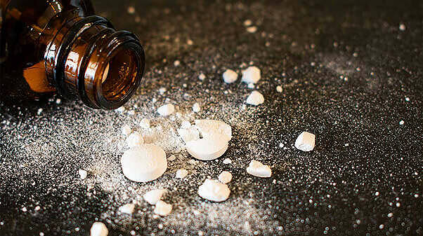Spilled white pills and powder next to an open brown pill bottle on dark surface, illustrating drug addiction dangers|Close-up of gloved hands writing on a medical chart with a urine sample cup nearby, representing documentation of Dilaudid use in a clinical setting