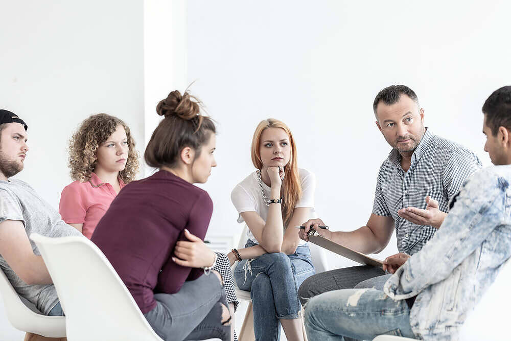School counselor explaining anger management techniques to a group of teenagers during a session.|Portrait of a solemn young woman with long dark hair, wearing a gray t-shirt, looking down with a troubled expression