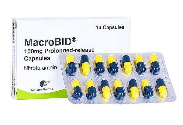 MacroBID 100mg prolonged-release capsules containing nitrofurantoin, showing blue and yellow capsules in blister pack|Woman contemplating the interaction between medication and alcohol, shown thinking about a cocktail and prescription pills|Close-up of a person's hand holding a glass of clear liquid, likely water or alcohol, with a blurred figure in a blue top in the background|Infographic showing a cross-section of a bacterial cell with labeled parts, illustrating four mechanisms of antibiotic action: disrupting cell wall synthesis, inhibiting RNA synthesis, inhibiting protein synthesis, and inhibiting DNA replication