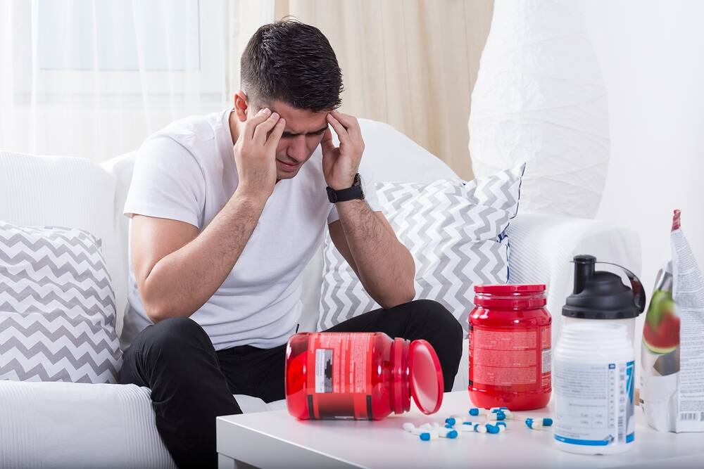 Distressed man holding his head, surrounded by supplement containers and pills on a coffee table|Close-up of a muscular arm lifting a heavy dumbbell, illustrating the effects of anabolic steroid use in bodybuilding and strength training