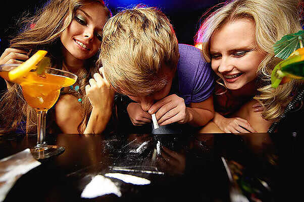 Young people using cocaine at a party, with alcoholic drinks visible, highlighting risks of party drug abuse|Close-up of hands exchanging small plastic bags containing various illegal substances, depicting the dangerous practice of sharing drugs|Graphic image with text "PARTY DRUGS" in bold letters against a dark blue background, highlighting the issue of recreational drug use|Broken purple pill capsule spilling white powder onto a wooden surface, with additional powder and rolled currency visible, illustrating prescription drug misuse|Several silver nitrous oxide canisters scattered on a weathered wooden surface, commonly used as a party drug known as 'laughing gas