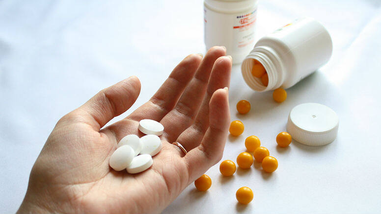 Hand holding white pills with orange pills scattered nearby and medication bottles in the background, illustrating prescription drug use and potential for addiction