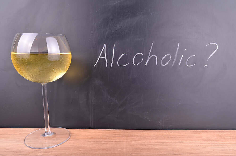 A glass of white wine next to a chalkboard with 'Alcoholic?' written on it, suggesting self-reflection on drinking habits|Infographic showing 14 units of alcohol per week represented as 6 pints of beer (4%) or 7 glasses of wine (175ml, 11.5%)|||sad woman sitting on sofa drinking wine two packs of desistal beside her|infographic-chart-showing-how-many-units-in-beer-wine-and-spirits-and-limits-for-men-and-women|image of desistal anticraving for people drinking too much wine with button to buy|Desistal anti alcohol craving packet for people drinking wine every day|Two silver pouches of Desistal Alcohol Recovery Pills with a shop now button below