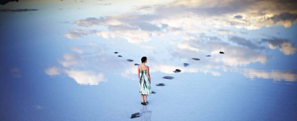 Woman in tie-dye dress walking on stepping stones across reflective water surface mirroring sky and clouds, symbolizing the path to healing and sobriety|Illustration of a woman experiencing hallucinations, imagining a conversation with a cartoon gnome, highlighting potential side effects of sleep medications like Ambien|Infographic showing 6 common symptoms of Ambien withdrawal: tremors, aches and pains, nausea, hyperventilation, panic attacks, and insomnia, illustrated with cartoon figures|Surreal image of a woman standing on water reflecting a cloudy sky, with stepping stones leading forward, symbolizing the path to recovery from addiction-induced hallucinations|Surreal image of a woman standing on a reflective blue surface with stepping stones leading forward, symbolizing the journey of addiction recovery against a cloudy sky background