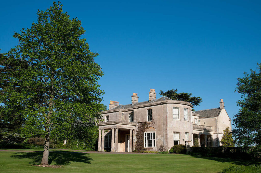 Elegant stone manor house with green lawns and trees, ideal for upscale rehab in Scotland|Picturesque stone building with Gothic architecture surrounded by lush gardens and colorful flowers, set against a backdrop of rolling hills, providing a tranquil setting for addiction recovery|Friendly group therapy session in a bright room with participants smiling and engaging, showcasing a welcoming and supportive environment for addiction recovery|Multi-generational group of four people hiking together on a nature trail, smiling and engaging in conversation, demonstrating the benefits of outdoor activities in addiction recovery|Middle-aged woman leading a diverse group therapy session with participants of various ages seated in a circle, in a bright room with plants, symbolizing supportive addiction recovery environment|Spacious and elegant living room in a rehab facility featuring a stone fireplace, comfortable sofas, large windows overlooking a garden, and modern decor, creating a serene and upscale environment for recovery