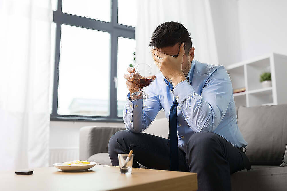 Stressed businessman holding wine glass, covering face with hand, displaying signs of alcohol addiction|Silhouette of a man holding a bottle of alcohol, surrounded by various bottles, symbolizing alcohol addiction.