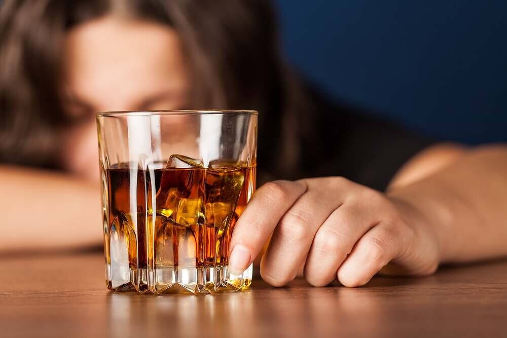 Close-up of a woman resting her head on a table next to a glass of whiskey, depicting alcohol abuse|Woman with dark hair slumped over table, holding a glass of red wine, depicting alcohol addiction and its effects|Infographic showing human body outline with labeled organs and corresponding symptoms of alcoholism, including effects on brain, heart, liver, stomach, and other organs|Detailed infographic showing a human silhouette drinking alcohol, with labeled organs and corresponding symptoms of alcoholism, including effects on brain, heart, liver, lungs, stomach, pancreas, and muscles|Woman's hands with red nail polish pouring whiskey from bottle into glass, depicting struggle with alcohol dependency|Man with head down on bar next to glass of whiskey, depicting effects of excessive alcohol consumption and potential addiction|Infographic showing a gauge depicting the spectrum of Alcohol Use Disorder from mild to severe, with explanatory text about AUD as a medical condition|Infographic showing a silhouette of a person drinking alcohol, with labeled illustrations of various organs and their corresponding symptoms and effects caused by alcoholism|Asphalt road with yellow arrow markings and text "ROAD TO RECOVERY" painted on the surface, symbolizing the path to addiction treatment and healing|Close-up of people sitting in a circle during a group therapy session, focusing on gesturing hands during discussion|Man with hands on head looking distressed, sitting at table with glass of whiskey and bottle in background, depicting struggle with alcohol addiction