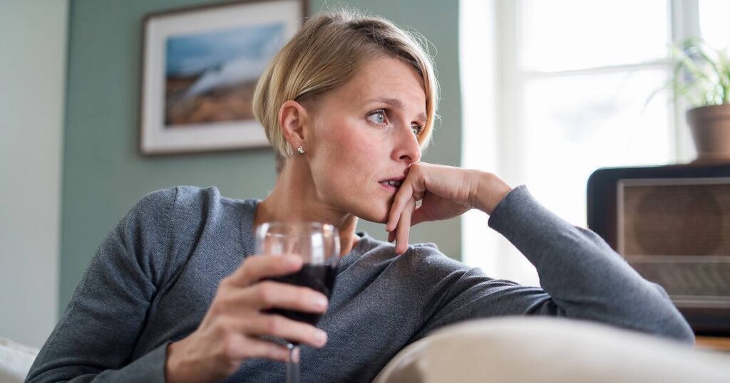 Concerned young blonde woman in gray sweater sitting alone, holding a glass of red wine while looking distressed and contemplative, suggesting alcohol dependency issues|Person with head in hands at bar counter, surrounded by alcohol shots and car keys, illustrating the dangers of drunk driving|Person slumped on wooden table with empty bottle and glass of alcohol, depicting the harmful effects of alcohol addiction|alcoholic drinking wine